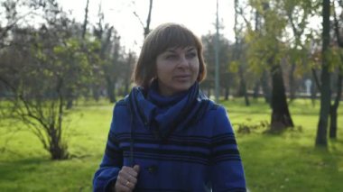 Sıska sarışın bir kadın, sonbaharda güneşli bir parka gidip gülümsüyor. Yavaş çekimde, güneşli bir yaz gününde, şık bir ceketle gezen ve gülümseyen, kısa saçlı, mutlu bir sarışın kadın manzarası harika.