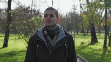 Cesur esmer çocuk, sonbaharda, güneşli bir sonbaharda, güneşli bir sonbahar gününde, güneşli bir parkta ayakta mutlu bir şekilde gülümseyen, spor ceketli, esmer bir çocuk görüntüsü içinde, ağır çekimde gülümsüyor. Harika görünüyor..
