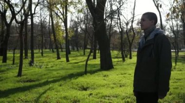 Neşeli genç bir adam güneşli bir parkta yürüyor ve sonbaharda yavaş çekimde gülümsüyor. Spor ceketli kel bir adamın güneşli bir sonbaharda güneşli bir parkta yürüyüşü ve gülüşü muhteşem..