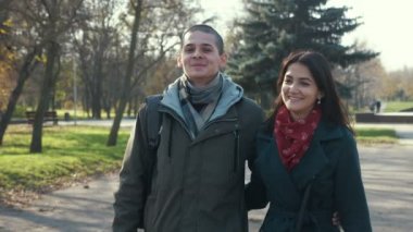 Neşeli esmer kız, neşeli erkek arkadaşıyla parkta ağır çekimde neşeli bir şekilde yürüyor. Genç ve mutlu bir esmer adamın uzun ve gevşek saçlarını sonbaharda yavaş çekimde güzel bir parkta gezdirmesini ve elini tutmasını keyifle seyrediyor..