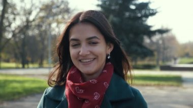 Neşeli esmer kız, güneşli bir parkta güneşli bir parkta mutlu bir şekilde gülümsüyor. Güzel bir parkta güzel bir esmer kız manzarası, uzun sarı saçları, kırmızı bir atkısı ve şık bir ceketi var. Güz mevsiminde güzel bir parkta neşeli bir şekilde gülümsüyor.