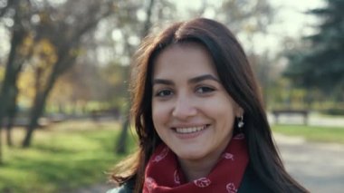 Güzel esmer kız, güneşli bir parkta, güzel bir parkta mutlu bir şekilde gülümsüyor. Hoş bir parkta, güzel, esmer, uzun, bol saçlı, kırmızı atkılı ve şık montlu bir kadının manzarası. Sonbaharda, resimli bir parkta mutlu bir şekilde durup gülümsüyor.