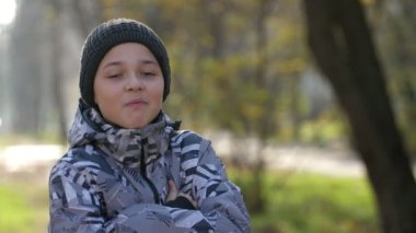Sonbaharda güneşli bir parkta ayakta duran ve gülümseyen kolları çapraz kollu neşeli çocuk. Parkta gülerek gülen siyah örgü şapkalı, spor ceketli 11 yaşında mutlu bir beyaz çocuğun neşeli portresi..