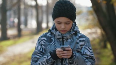Zeki çocuk dışarıda duruyor ve sonbaharda dışarıda video oyunları oynuyor. 11 yaşında örgü şapkalı ve ceketli bir çocuğun güneşli bir sonbaharda parkta video oyunu oynadığı harika bir manzara..