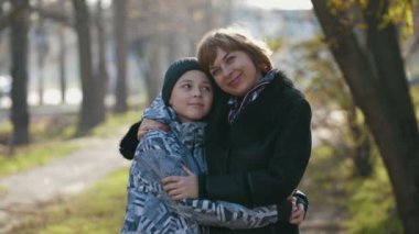 Mutlu sarışın anne, güzel bir sonbahar gününde küçük oğluna sarılıyor. Neşeli bir aile manzarası harika. Zarif bir ceketle gülümseyen sarışın anne, sonbaharda dışarıda omzuna yaslanan 11 yaşındaki oğlunu kucaklıyor. Güzel görünüyor..