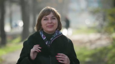 Güzel sarışın kadın, sonbaharda açık havada şık bir paltoyla ayakta duruyor ve gülümsüyor. Neşeli bir bokeh portresi. Şık montlu, renkli bir atkısı olan neşeli, sarışın bir kadın..