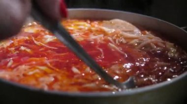 Kadın eli kaşığı tutuyor ve metal bir güveçte kaynamış borş taşıyor. Metalik bir kaşığı tutan bir kadının neşeli yakın çekimi ve metalik bir tencerede kaynamış kırmızı ve sarı Ukrayna borşunu karıştırıyor. İyimser ve lezzetli görünüyor..