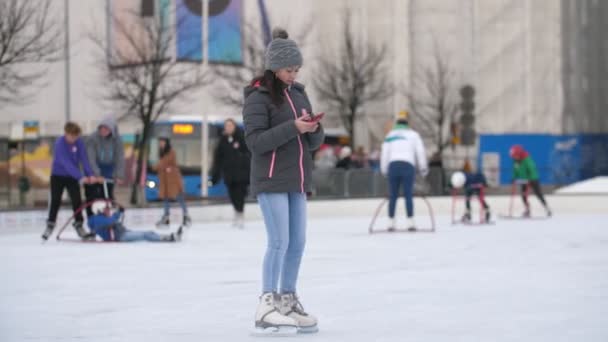 フィンランド ヘルシンキ 2019年12月12日 スレンダーな女の子がスマートフォンを手にして 冬にはゆっくりとした動きでヘルシンキのストリートアイスリンクで楽しくスケートをしています 彼女は幸せそうだ — ストック動画