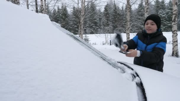 フィンランドの雪に覆われた森の中で長いほうきで車のフロントガラスを掃除する陽気な少年フィンランドの冬に絵のように美しい松林の中で重い雪の結晶で覆われたフロントガラスを拭くとクリーニング11歳の少年の素晴らしい景色 — ストック動画