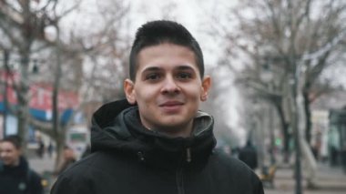 Siyah ceketli neşeli esmer adam ağır çekimde ayakta duruyor ve gülümsüyor. Siyah kapüşonlu mutlu, genç bir adamın iyimser portresi. Sonbaharda ağaçlarla dolu bir sokakta dikiliyor ve gülümsüyor.. 