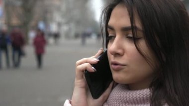 Güzel esmer kız, arkadaşlarıyla telefonda ağır çekimde ağır çekimde dışarıda. Uzun, gevşek saçlı, gergin bir esmer kızın ağır çekimde ağaçlarla dolu gri bir sokakta arkadaşıyla telefonda küfür edişinin duygusal görüntüsü.