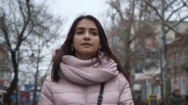 Neşeli ve hoş bir kız gri bir caddede ağır çekimde geziniyor. Uzun, gevşek saçlı, parlak gözlü, pembe ceketli genç bir kızın heyecan verici manzarası. Sonbaharda bir caddede ve sokakta ağır çekimde yürüyor.