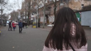 Uzun saçlı, zayıf bir kız yavaş çekimde sokakta yürüyor ve geri dönüyor. Uzun, gevşek saçlı, zayıf esmer bir kızın arka görünüşü sonbaharda insanlarla birlikte sokakta dönüp duruyor..