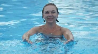 Bikinili mutlu sarışın kadın mavi suyu yavaş çekimde eğlendiriyor ve okşuyor. Bikinili gülümseyen sarışın kadın ayakta, düz elleriyle hareket ediyor ve mavi suyu yavaş çekimde yüzme havuzunda okşuyor..