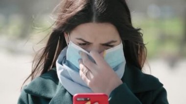 Hijyenik maske takmış endişeli genç bir kadın ilkbaharda güneşli bir bankta telefonunda enidemik yayılmış Coronavirus 'a karşı hijyenik maske takan endişeli esmer bir kızın rıhtımda Covid-19 hakkında bilgi arıyor.