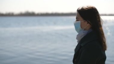 Güzel esmer kız ilkbaharda gün batımında parlayan Dnipro nehrine bakıyor. Coronavirus 'a karşı hijyenik maske takmış romantik esmer bir kadının ilkbaharda gün batımında Dinyeper sularına bakması için ilham veriyor.