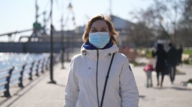 Hijyenik maske takan zarif sarışın kadın yavaş çekimde Dnipro rıhtımında yürüyor. Beyaz ceketli şık sarışın bir kadın ve Coronavirus 'a karşı koruyucu bir maske takıyor. Güneşli bir baharda güneşli bir baharda islerde sokak lambalarıyla rıhtıma gidiyor.