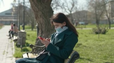 Hijyenik maskeli zarif bir kız, bahar parkında otururken akıllı telefonunu çeviriyor. Uzun saçlı, hijyenik maske takan, koronavirüsüne karşı iyimser bir kız. Ve bahar parkındaki bir bankta oturan akıllı telefonunu çeviriyor.