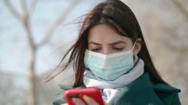 Hijyenik maskeli güzel bir kız, ilkbaharda açık havada akıllı telefonunu arıyor. Uzun saçlı, hijyenik bir maske takan, akıllı telefonunu arayan, koronavirüsü görmezden gelen ve bahar havasının tadını çıkaran çekici esmer bir kız.