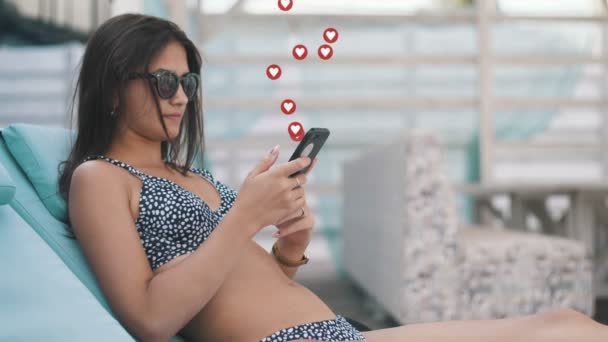 海のリゾートでリクライナーに座っているビキニとサングラスの魅力的な女の子の素晴らしい景色と彼女の携帯電話上のソーシャルネットワークから多くの同類を取得します ユーザーインターフェイスアニメーション — ストック動画