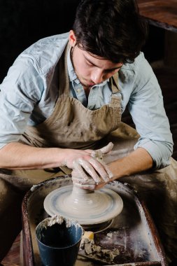 çanak çömlek, atölye, Seramik Sanat kavramı - potter's tekerlekli el ile ham kil ile çalışan adam bir erkek esmer heykeltraşlık araçlarıyla, önlük ve bir gömlek ana ahşap masa yakınındaki bir mutfak eşyaları