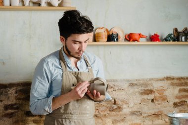 Potter, atölye, Seramik Sanat kavramı - holding Fransız Kupası ve ürün, erkek usta bir lekeli kil önlük giymiş genç dikkatle inceliyor, Usta'nın ellerini tutarak çiğ sürahi