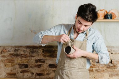 Potter, atölye, Seramik Sanat kavramı - holding Fransız Kupası ve ürün, erkek usta bir lekeli kil önlük giymiş genç dikkatle inceliyor, Usta'nın ellerini tutarak çiğ sürahi