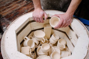 pottery, workshop, ceramics art concept - closeup on male hands gently puts the jug in the kiln, top view of electric oven for further roasting of unfinished clay products, cups and unbaked utensils clipart