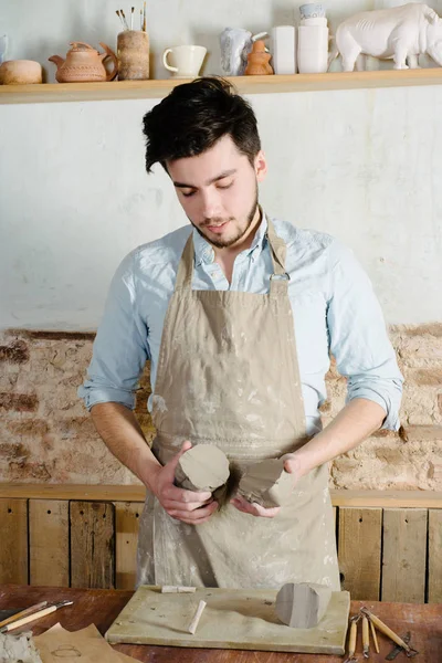 potter, workshop, ceramics art concept - standing young brunette man dressed in an apron, male hands knead the fireclay, a ceramist with raw materials on wooden table with sculpting tools set