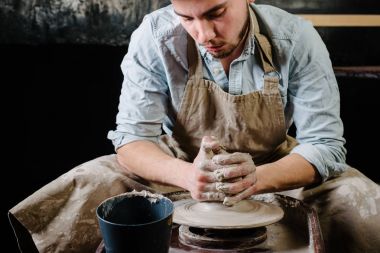 çanak çömlek, atölye, Seramik Sanat kavramı - potter's tekerlekli el ile ham kil ile çalışan adam bir erkek esmer heykeltraşlık araçlarıyla, önlük ve bir gömlek ana ahşap masa yakınındaki bir mutfak eşyaları