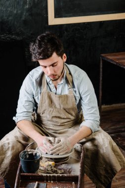 çanak çömlek, atölye, Seramik Sanat kavramı - potter's tekerlekli el ile ham kil ile çalışan adam bir erkek esmer heykeltraşlık araçlarıyla, önlük ve bir gömlek ana ahşap masa yakınındaki bir mutfak eşyaları