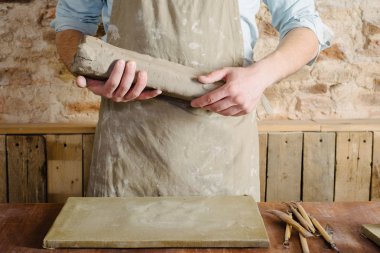 Potter, atölye, Seramik Sanat kavramı - genç esmer adam ayakta bir önlük giymiş, erkek eller yoğurmak şamot hammadde heykel araçlar kümesi ile ahşap tablo ile bir ustası