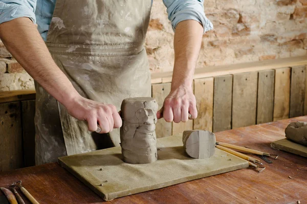 potter, clay workshop, ceramics art concept - male cuts raw clay with the string cutter, closeup on hands of standing man dressed in an apron, ceramist work over wooden table with sculpting tools set