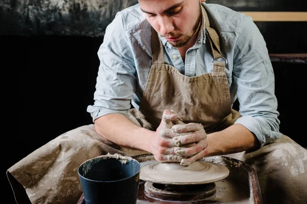 çanak çömlek, atölye, Seramik Sanat kavramı - potter's tekerlekli el ile ham kil ile çalışan adam bir erkek esmer heykeltraşlık araçlarıyla, önlük ve bir gömlek ana ahşap masa yakınındaki bir mutfak eşyaları