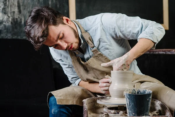 çanak çömlek, atölye, Seramik Sanat kavramı - potter's tekerlekli el ile ham kil ile çalışan adam bir erkek esmer heykeltraşlık araçlarıyla, önlük ve bir gömlek ana ahşap masa yakınındaki bir mutfak eşyaları