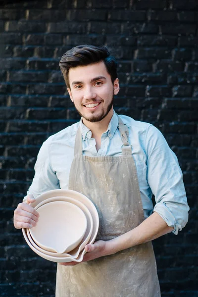 potter, utensils, ceramics art concept - smiling male ceramist in the apron shows samples of clay products, craftsman holding new finished bowls, happy master on background of dark gray brick wall