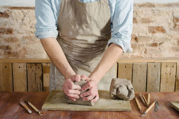 Potter, workshop, ceramics art concept - standing young brunette man dressed in an apron, male hands knead the fireclay, a ceramist with raw materials on wooden table with sculpting tools set — Stock Photo, Image