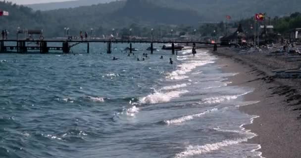 Vista de las olas en la línea de la costa del mar con gente y barcos amarrados a muelle en el día de verano — Vídeo de stock