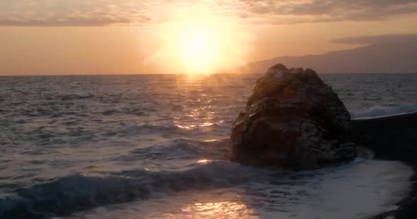 Vedere a valurilor laminate pe coasta mării cu o stâncă uriașă la apusul soarelui — Videoclip de stoc