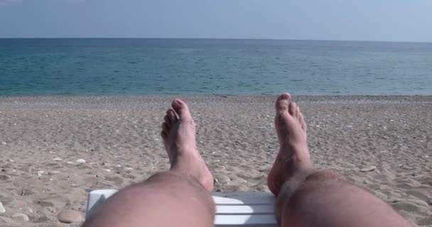 Man benen op zonnebank, uitzicht op het strand en de zee — Stockvideo