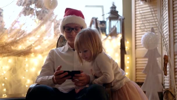 Little Cute Girl and Her Elder Brother in Santa Hat Having Fun With Mobile Phone in the Rocking Chair — Stock Video