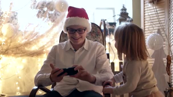 Roztomilá holčička a její starší bratr v Santa Hat baví s mobilním telefonem v houpacím křesle — Stock video