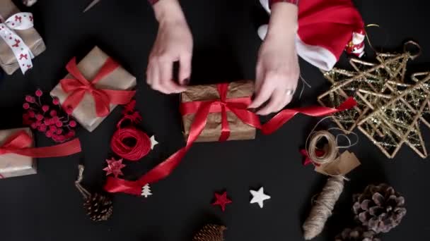 Mani in alto Avvolgendo regali di Natale su sfondo nero dall'alto. Regalo avvolto in carta marrone e nastro bianco legato — Video Stock
