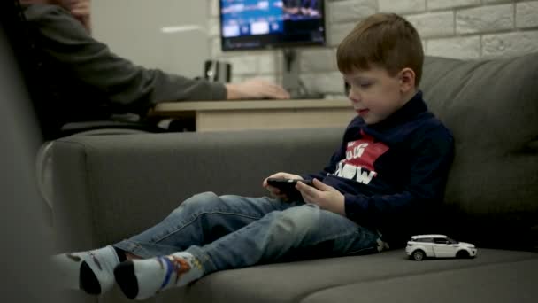 Menino jovem sentado no sofá e jogando jogos no telefone inteligente . — Vídeo de Stock