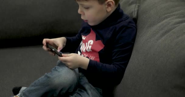 Kleiner Junge sitzt auf der Couch und spielt Spiele auf dem Smartphone. — Stockvideo