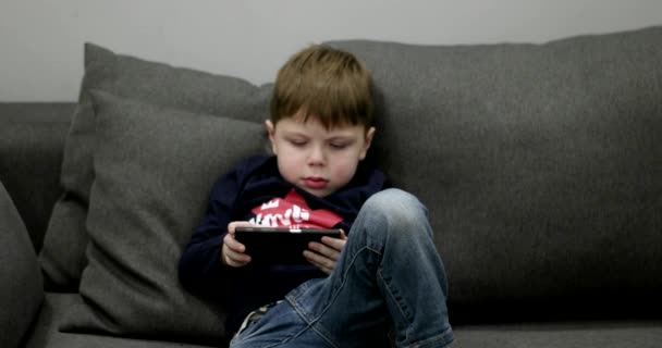 Menino jovem sentado no sofá e jogando jogos no telefone inteligente . — Vídeo de Stock