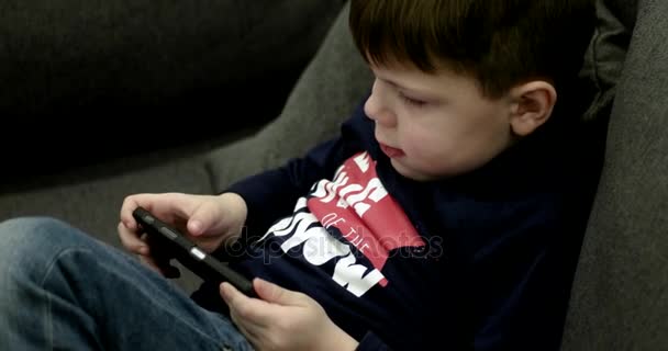 Niño sentado en el sofá y jugando juegos en el teléfono inteligente . — Vídeos de Stock