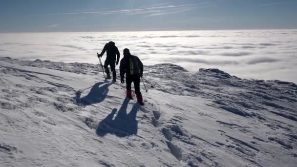 Viajantes com mochilas fazem uma caminhada de inverno acima das nuvens com vista para os picos distantes por uma costa do mar da nuvem — Vídeo de Stock