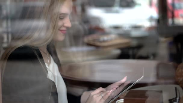 Fiatal üzleti nő ül egy asztalnál, ivott kávét, teát és gépelés-ra tabletta pc egy kávézóban, étteremben tabletta számítógép segítségével
