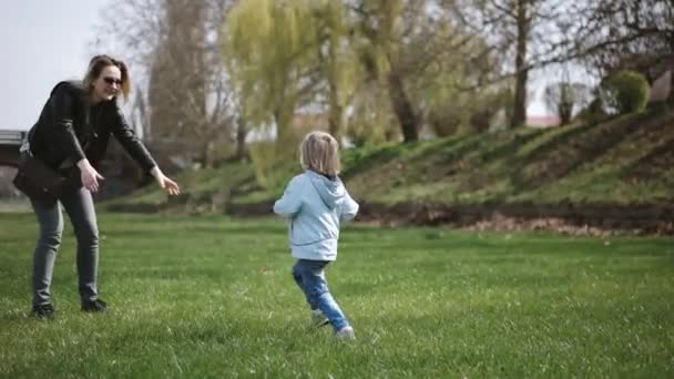 Copilul aleargă în mâinile mamei pentru a o îmbrățișa și a se roti în jurul ei. Familia se distrează în parc pe o iarbă verde. Slow Motion — Videoclip de stoc